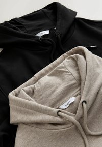 To små hættetrøjer, en sort og en lysegrå, lavet af blødt bomuldsstof. Begge har snore, kængurulommer og et mærke indeni.