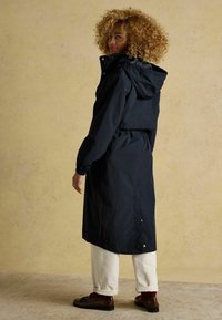 Jachetă impermeabilă de marina cu glugă, talie strânsă și mâneci fără degete; are un siluet lung și nasturi cu accente aurii la tiv.