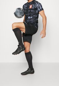 Camiseta de fútbol negra de manga corta con diseño estampado y logo rojo, combinada con pantalones cortos negros, medias largas negras y zapatillas de fútbol negras. Balón de fútbol en alto.