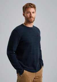 Marineblauer Langarmpullover mit Rundhalsausschnitt und gerippter Textur, versehen mit einem Brustlogo-Patch. Getragen mit tanfarbenen Cargo-Hosen.