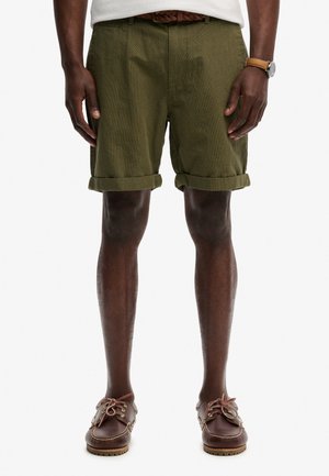 Osoba ubrana w oliwkowo zielone, podwinięte szorty, brązowy pleciony pasek, brązowe skórzane buty i zegarek na lewej ręce, stojąca z rozluźnionymi rękami.