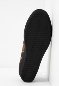 Semelle d'une chaussure en caoutchouc noire avec un motif de grip texturé et un tissu à imprimé léopard sur le côté, positionnée verticalement contre une surface blanche.