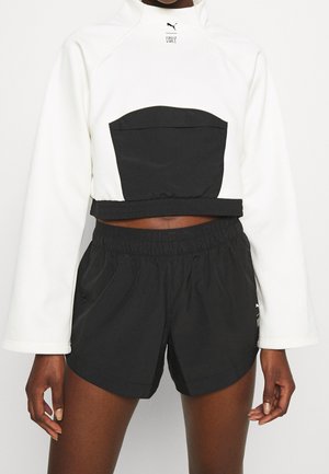 Personne portant un haut court à manches longues blanc avec une poche avant noire et un short de sport noir à taille élastique, debout devant un fond blanc.