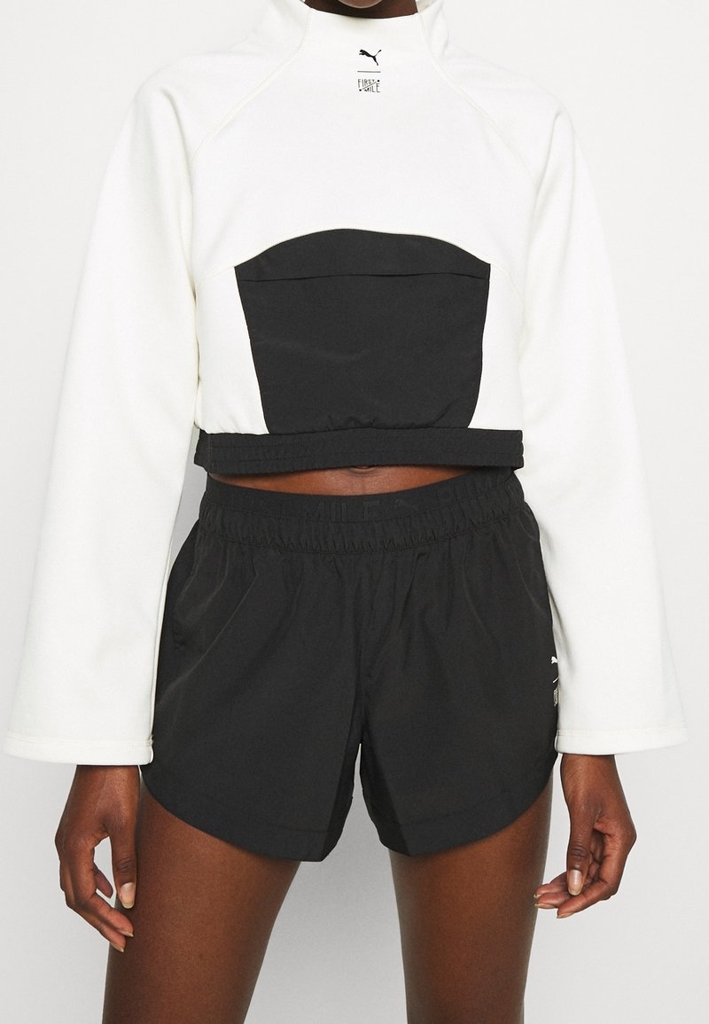 Personne portant un haut court à manches longues blanc avec une poche avant noire et un short de sport noir à taille élastique, debout devant un fond blanc.