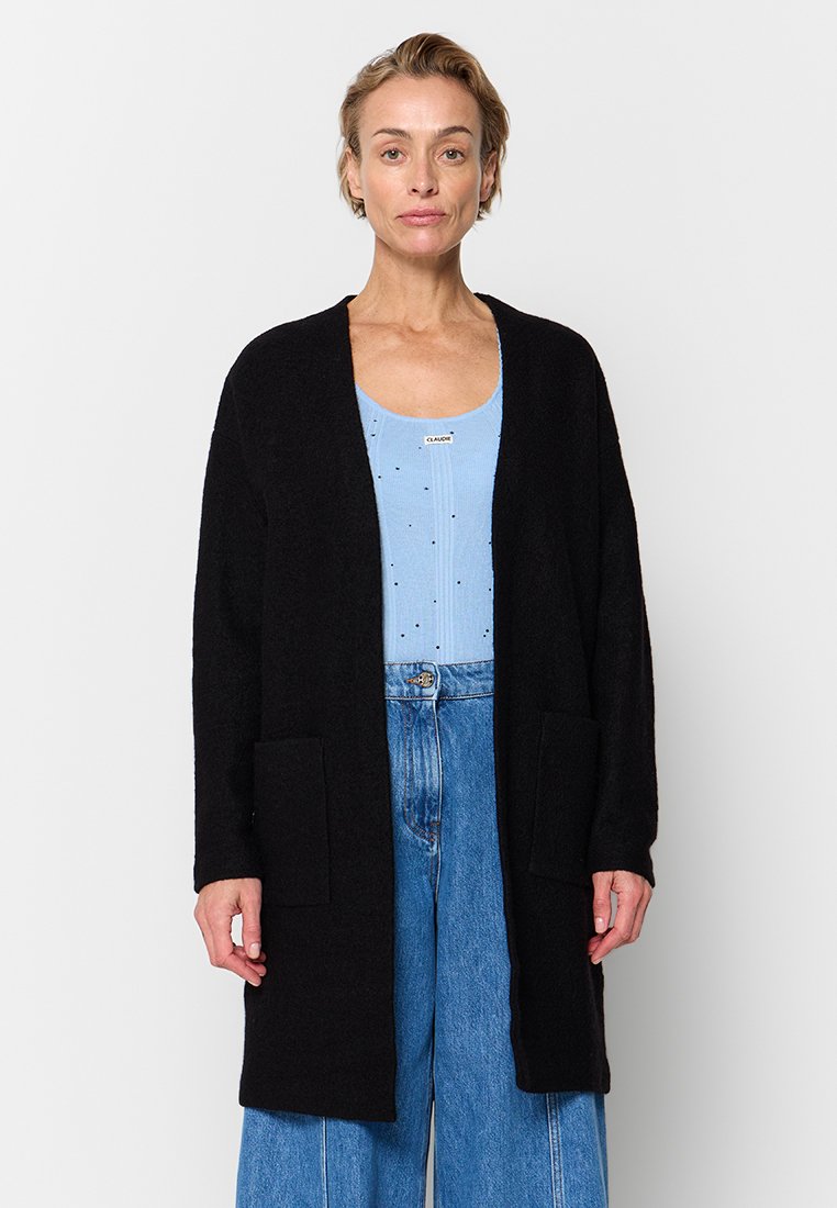 Zwarte cardigan met lange mouwen, open voorkant, twee voorzakken en een zachte textuur, gedragen over een lichtblauw topje en losse denimbroek.