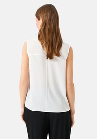 Weißes ärmelloses Oberteil mit abgerundetem Saum, subtiler Struktur und einer Passe über dem oberen Rücken. Kombiniert mit schwarzen Hosen.