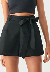 Svarta linnebyxor med bred midjeband och snörning fram. Har sidofickor och en lös passform, med en slät, matt textur.