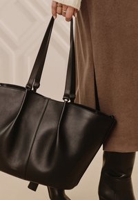 Czarna skórzana torba na ramię z podwójnymi uchwytami, o gładkiej fakturze i subtelnym plisowanym wzorze. Nosi się ją w zestawieniu z brązowym strojem i kozakami za kolano.