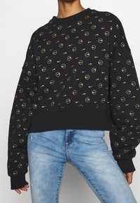 Svart sweatshirt med ett guldigt cirkulärt mönster och Nike-logotyper, rundad halsringning och ribbade ärmslut, kombinerad med blå utslitna jeans.