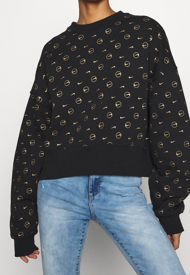 Svart sweatshirt med ett guldigt cirkulärt mönster och Nike-logotyper, rundad halsringning och ribbade ärmslut, kombinerad med blå utslitna jeans.