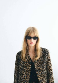 Zlatý blazer s leopardím potiskem a černými knoflíky, spárovaný s černým topem. Modelka nosí úzké černé sluneční brýle a má rovné světle hnědé vlasy.