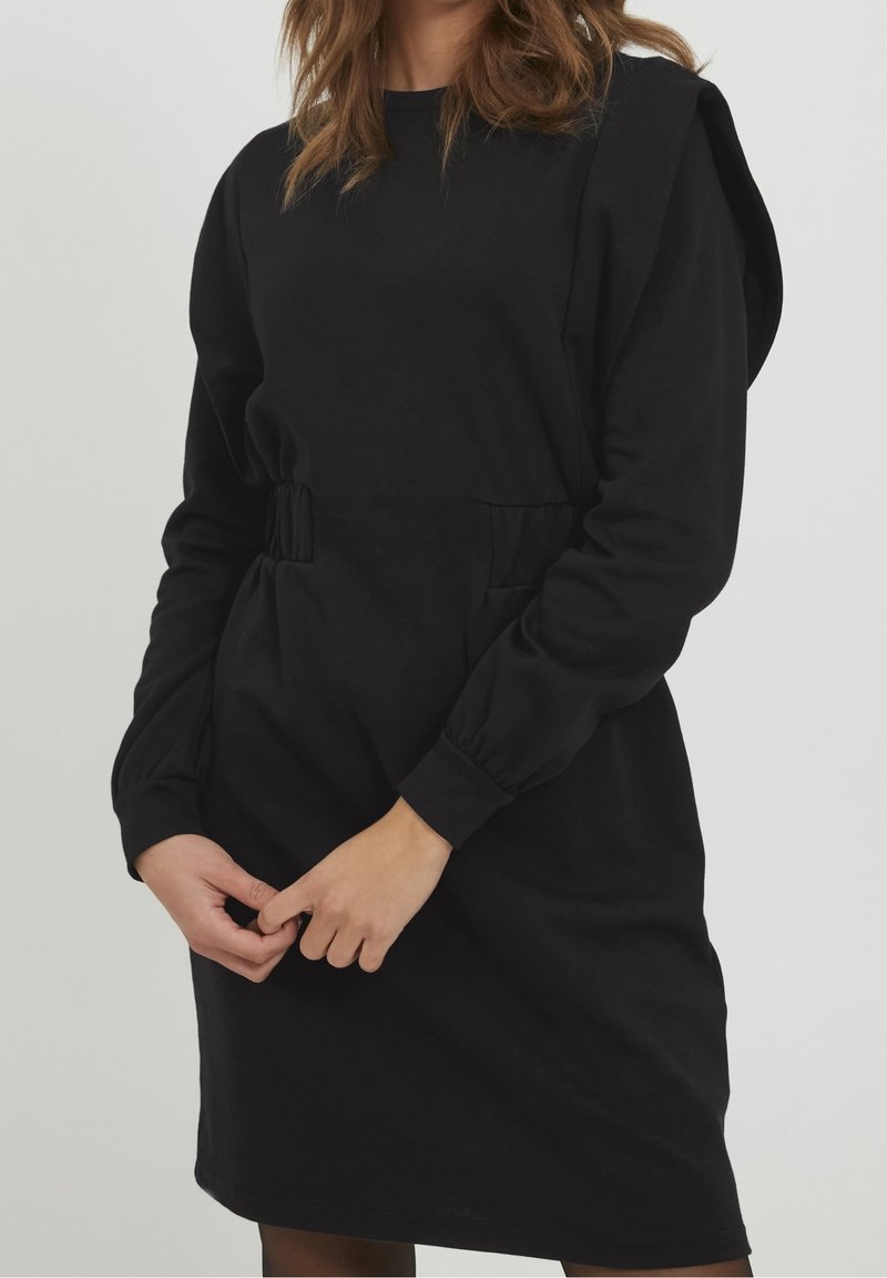 Robe noire en tissu mélange de coton, dotée d'un col rond, de manches longues, d'une taille élastique et de poches latérales. Texture lisse et coupe décontractée.