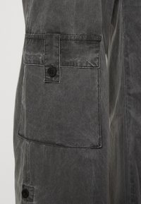 Detailní záběr na šedou bundu, která zdůrazňuje velkou kapsu s černým knoflíkem a texturovanou tkaninou, s důrazem na její materiál a detaily.