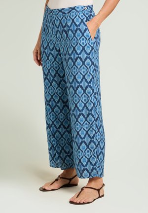Frau trägt blaue, weit geschnittene Hose mit Seitentaschen und braune flache Sandalen, steht vor hellem Hintergrund.