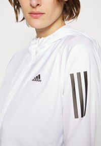 Valkoinen vetoketjullinen takki, joka on valmistettu kevyestä materiaalista ja siinä on teksturoitu pinta. Takissa on musta Adidas-logo ja kolme mustaa raitaa hihassa.