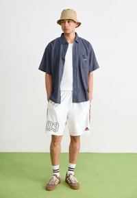 Rutigt kortärmat skjorta i blått, vita träningsshorts med tryck av '03', beige bucket hat och bruna snörskor med vita sulor.