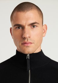 Schwarzer Reißverschluss-Pullover mit hohem Kragen, gerippter Textur und silbernem Reißverschluss. Glattes Material und eine enganliegende Passform am Hals.