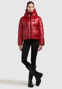 Vrouw in een glanzende rode pufferjack met capuchon, zwarte leggings en zwarte kniehoge laarzen, staande tegen een egale lichte achtergrond.