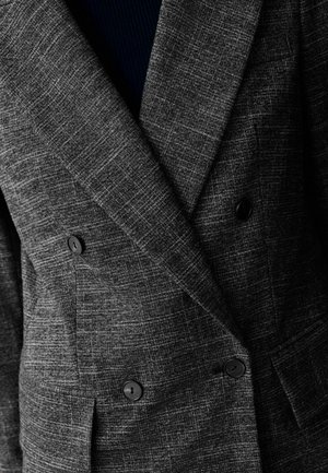 Šedá, texturovaná dvouřadá bunda s houndstooth vzorem, černými knoflíky a širokými klopami. Vnitřní vrstva viditelná v tmavě modré barvě.