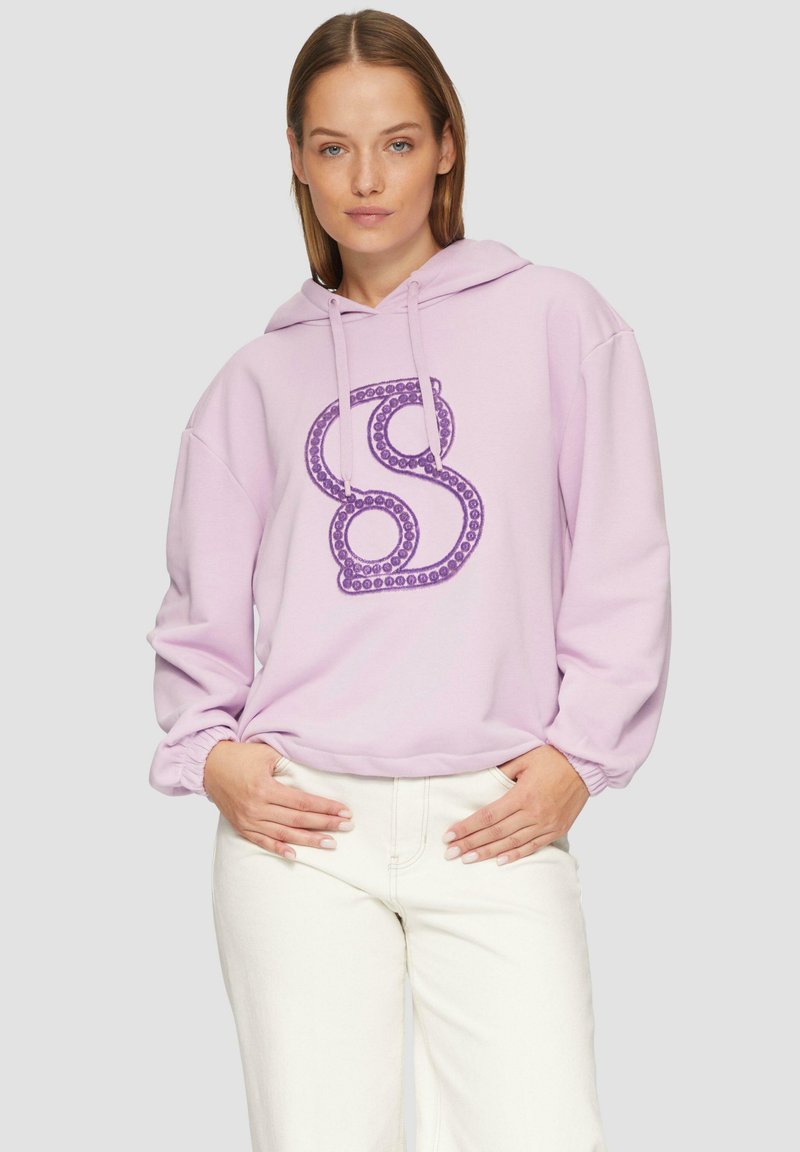 Felpa viola con una grande "S" decorativa in finitura lucida, caratterizzata da un cappuccio con coulisse e polsini arricciati, indossata sopra pantaloni bianchi.