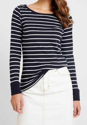 Frau trägt ein marineblau-weiß gestreiftes Langarmshirt mit weißem Rock, mit teilweise über der Schulter sichtbarem Haar.