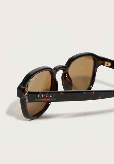Lunettes de soleil de style écaille avec des verres bruns, comportant le logo "GUCCI" et un détail de bande rouge-vert sur la branche.