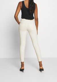 Žena stojící zády, oblečená v bílých úzkých džínách, černém bezrukávovém topu a černých sandálech na podpatku na bílém pozadí.