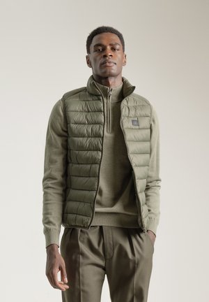 Chaleco de plumas verde oliva con un patrón acolchado, llevado sobre un suéter a juego, combinado con pantalones oliva, que cuentan con un cuello con cremallera y un diseño minimalista.