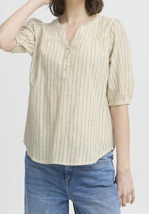 Femme portant un chemisier beige à rayures verticales avec des manches bouffantes trois-quarts et un jean bleu, posant devant un fond uni.