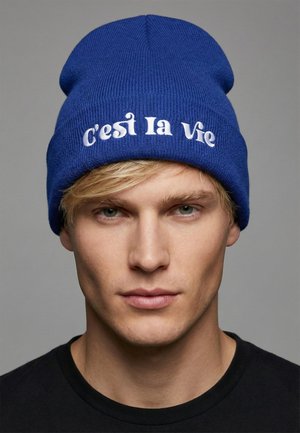 Blue knit beanie with embroidered white text reading "C'est la vie." The hat features a snug fit and a soft, textured fabric.