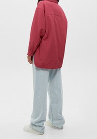 Chemise oversize à manches longues de couleur rose profond, en tissu fin, avec une couture de yoke au dos, associée à un jean large bleu clair et des chaussures blanches.