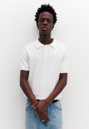 Ung mand med mørkt, krøllet hår iført en hvid kortærmet polo og lyseblå jeans, stående foran en hvid baggrund.