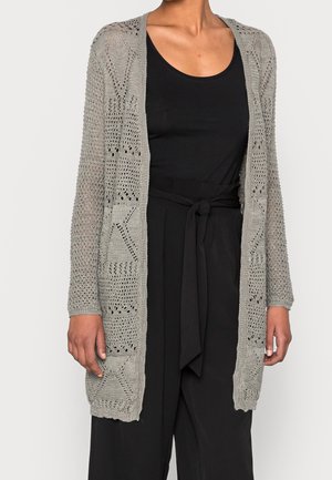 Donna che indossa un cardigan lungo in maglia aperta grigio chiaro sopra una canotta nera senza maniche e pantaloni neri a vita alta con cintura annodata.