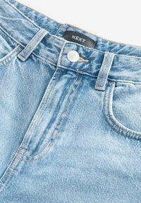Jean en denim bleu clair en gros plan montrant un bouton en métal, des passants de ceinture, une poche avant et une étiquette de marque NEXT noire sur la ceinture.
