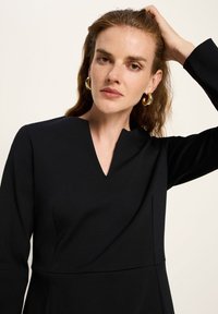 Frau mit heller Haut und welligem Haar, die goldene Creolen trägt und ein schwarzes langes Kleid mit V-Ausschnitt hat, während sie mit der Hand ihr Haar berührt.