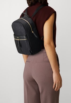 Femme portant un haut marron sans manches et un pantalon mauve avec un sac à dos noir orné de fermetures éclair dorées sur le dos.