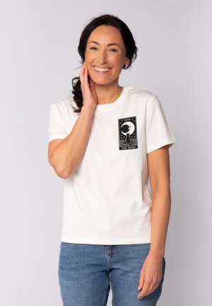 T-shirt en coton blanc avec un graphisme noir représentant une lune et des étoiles. Manches courtes, col rond et coupe décontractée. Porté avec un jean bleu.