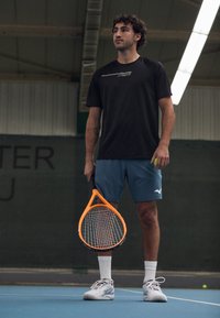 Mann i svart skjorte og blå shorts står på innendørs tennisbane og holder en oransje tennisracket og grønn ball.