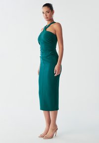 Vestido verde esmeralda elegante con diseño de un solo hombro y detalle fruncido. Largo hasta media pantorrilla, silueta ajustada, textura suave y discretos acentos de costuras.