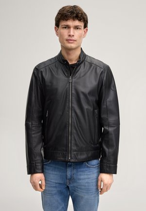 Giovane uomo con giacca di pelle nera con cerniera e jeans blu, in piedi contro uno sfondo chiaro e uniforme.