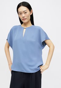 Zila īsām piedurknēm blūze ar atvērumu pie kakla, izgatavota no mīkstas auduma. Ir minimālistiska dizaina un smalka spīduma, ko papildina sudraba akcenti.