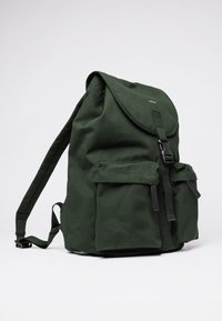 Zaino in tela verde scuro con chiusura a pattina, due tasche frontali, spallacci regolabili e dettagli in metallo nero. Superficie in tessuto testurizzato.