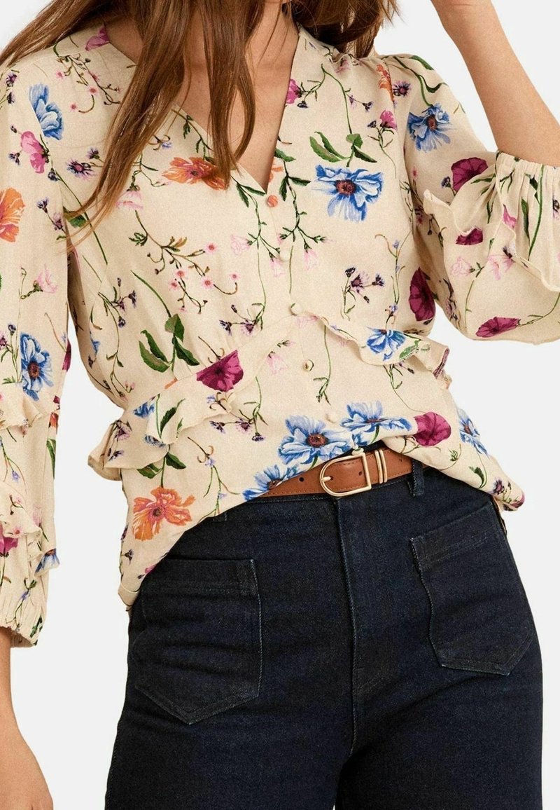 Femme portant un chemisier beige à fleurs bleues et roses, associé à un jean en denim foncé et une ceinture marron avec une boucle dorée.