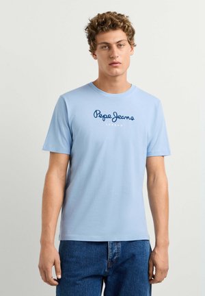 Joven con pelo rizado que lleva una camiseta azul claro de Pepe Jeans London y vaqueros azules sobre un fondo liso.