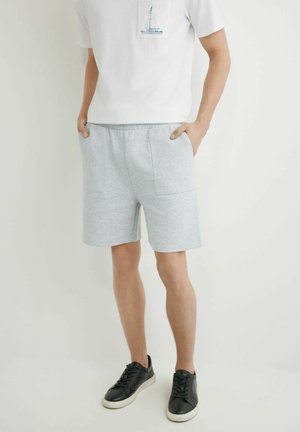 Mężczyzna noszący jasnoszorty do kolan z dużymi przednimi kieszeniami, białą koszulkę z nadrukiem żaglówki oraz czarne trampki z białymi podeszwami.