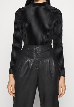 Kvinde iført en sort ribbet langærmet top, der er stukket ned i sorte læderbukser med høj talje, forplissering foran og knaplukning.