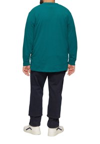 Türkises langärmeliges Shirt aus Baumwolle, kombiniert mit dunkelblauen Hosen und weißen Sneakers mit schwarzen Akzenten, von hinten betrachtet.