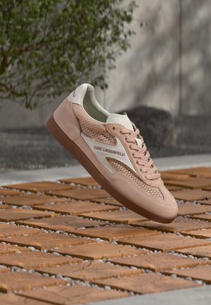 Baskets Karl Lagerfeld rose clair avec panneaux en maille et accents blancs, flottant au-dessus de carreaux en bois en extérieur avec une verdure floue en arrière-plan.