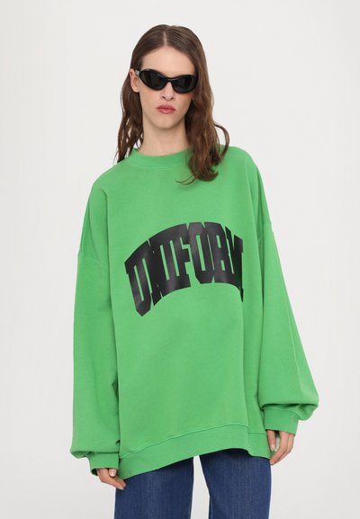 Grønn oversized sweatshirt med lange ermer, med en dristig svart logo på forsiden. Modellen har på seg svarte solbriller. Uformell design.