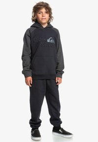 Conjunto de sudadera y pantalones de chándal en azul marino oscuro. La sudadera tiene un bolsillo tipo canguro y un logo texturizado. La tela parece suave; incluye acentos grises en las mangas.
